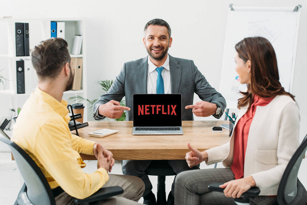 advisor pointing with fingers at laptop with netflix website on screen near man and woman thumbing up in office - Foto, Imagen
