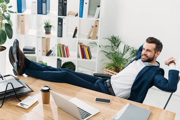 uomo d'affari in abito blu seduto con le gambe sul tavolo e sorridente in ufficio
 - Foto, immagini