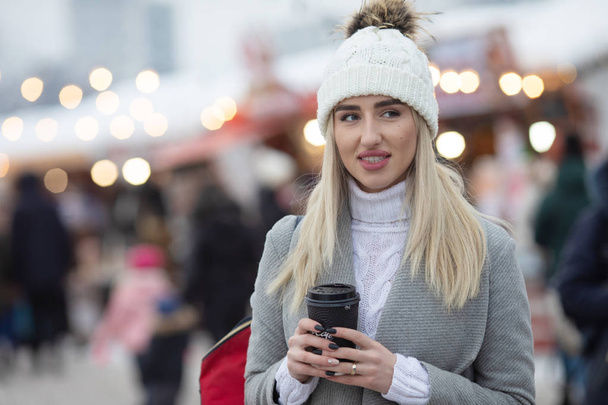 Outdoors lifestyle fashion portrait of pretty blonde girl walking on the holiday city in blizzard. Smiling, drinking coffee. Christmas mood. - Foto, afbeelding