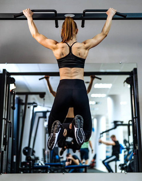 Muscular, athletic girl, bodybuilder in sportswear pulling up on a horizontal bar in front of the mirror at gym. Back muscles of a sporty girl tightening and looking at herself in the mirror. - Foto, afbeelding