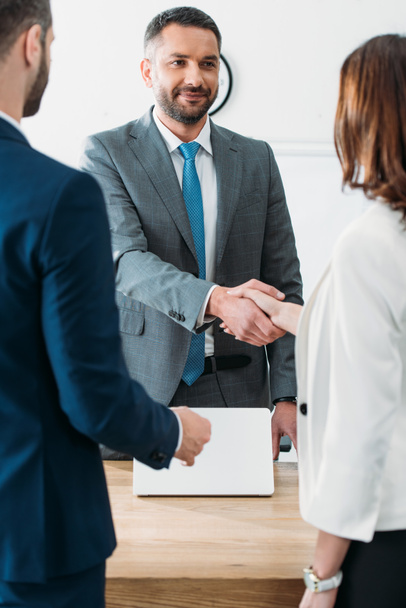 selective focus of handsome advisor in suit and investors shaking hands at workplace   - Zdjęcie, obraz