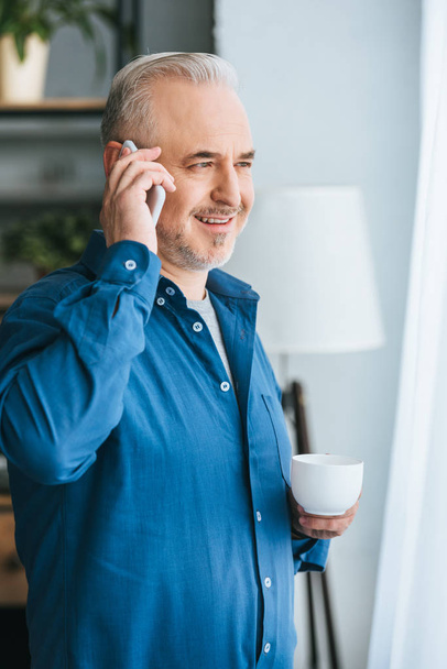 cheerful man holding cup and smiling while talking on smartphone - Photo, Image