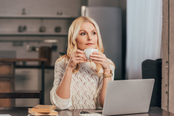 привлекательная задумчивая женщина держит чашку с напитком возле ноутбука
 - Фото, изображение