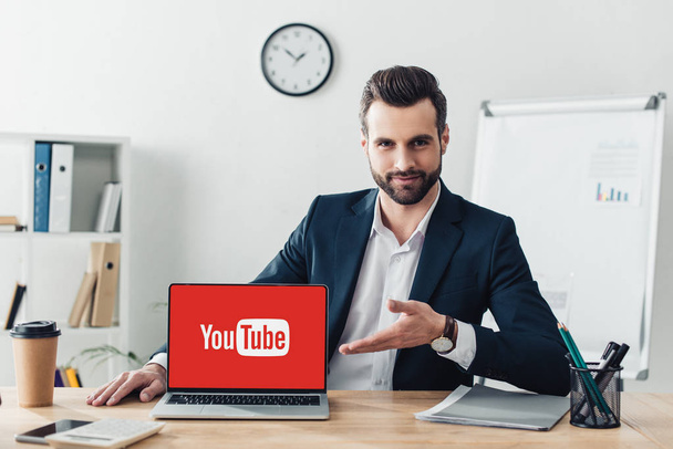 handsome advisor in suit pointing with fingers at laptop with youtube website on screen at office  - Foto, Bild