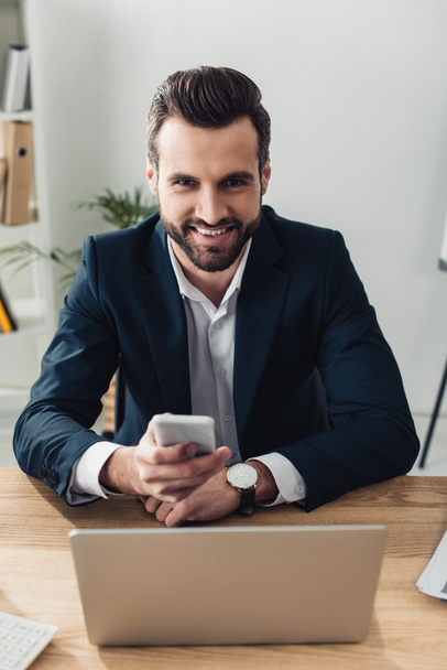 asesor sonriente y guapo en traje usando smartphone en el lugar de trabajo
  - Foto, Imagen