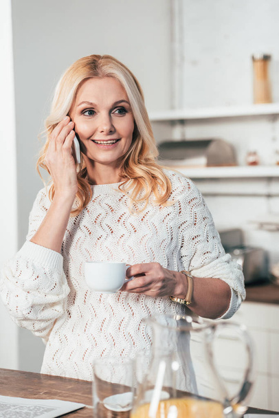 cheerful woman talking on smartphone while holding cup with drink in kitchen  - Fotografie, Obrázek