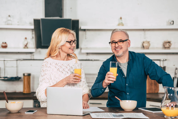 cheerful woman looking at husband in eye glasses holding glass with orange juice near laptop - Foto, Imagen