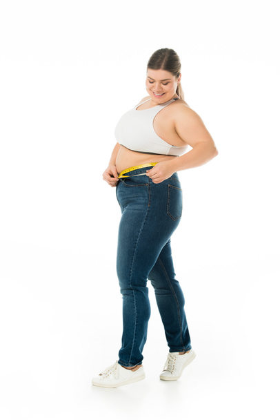 mulher com sobrepeso sorridente em jeans medindo a cintura com fita métrica isolada em branco, conceito de positividade corporal
 - Foto, Imagem