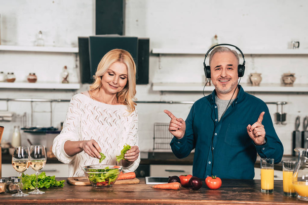 cheerful woman preparing food near happy husband listening music in headphones in kitchen - Foto, imagen