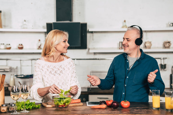 Szczęśliwa kobieta przygotowywania żywności w pobliżu mąż szczęśliwy słuchania muzyki w słuchawkach w kuchni - Zdjęcie, obraz