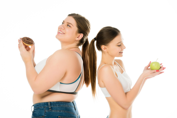 happy slim and overweight girls standing back to back together with doughnut and green apple isolated on white - Photo, Image
