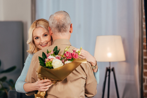 cheerful woman smiling while hugging husband and holding flowers and gift box in hands - Photo, image
