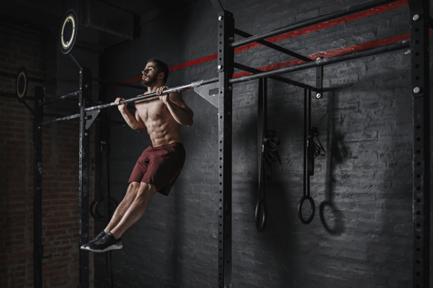 Crossfit athlete doing pull-ups at the gym. Practicing calisthenics. Handsome man doing functional training. - 写真・画像
