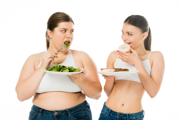 λεπτή γυναίκα τρώει ντόνατς και υπέρβαρη γυναίκα τρώει φύλλα πράσινο σπανάκι ενώ ψάχνει σε κάθε άλλο απομονωθεί σε λευκό - Φωτογραφία, εικόνα