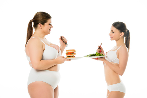 schlanke Frau in Unterwäsche mit Teller mit grünen Spinatblättern und Blick auf übergewichtige Frau mit Teller mit Burger isoliert auf weiß - Foto, Bild