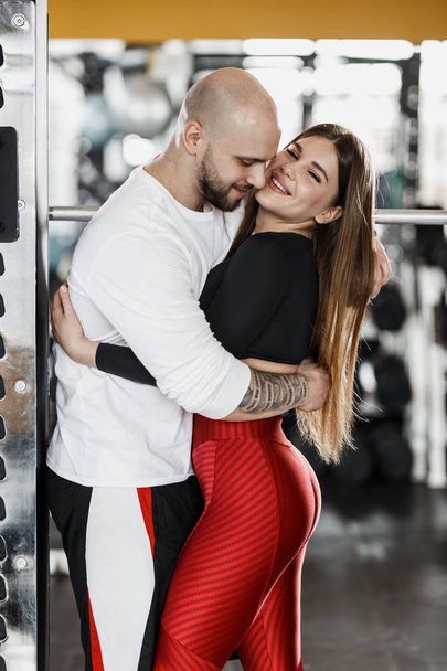 Romantic happy athletic couple. Strong man and slender beautiful girl are hugging in the modern gym next to the sport equipment - Zdjęcie, obraz