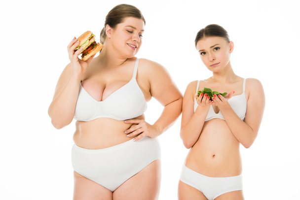 slim upset woman in underwear holding green spinach leaves while overweight happy woman eating burger isolated on white - Zdjęcie, obraz