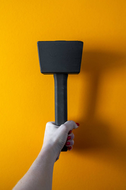 hand of a young woman holding a black wooden hammer, nails covered with red varnish, bright yellow background, close-up, vertical shot - Foto, Imagem