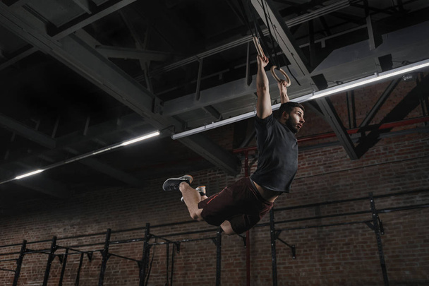 Young athlete doing pull-ups on gymnastic rings at cross fit gym. Handsome man practicing muscle ups swinging workout exercise. - Foto, immagini