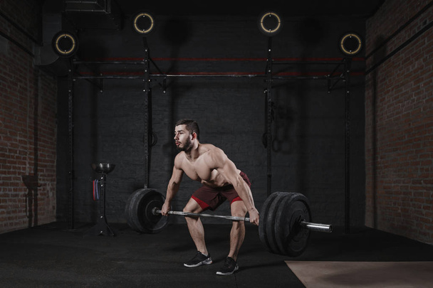 Young shirtless athlete lifting heavy barbell at crossfit gym. Handsome man practicing powerlifting deadlift exercise. - Photo, Image