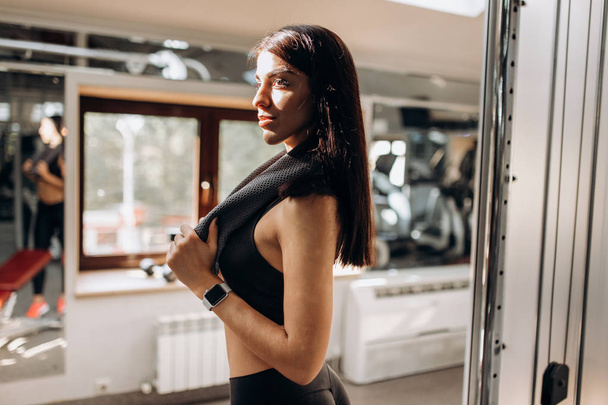 Beautiful athletic girl dressed in black sports top and tights stands in the gym with the towel on her shoulders - Valokuva, kuva