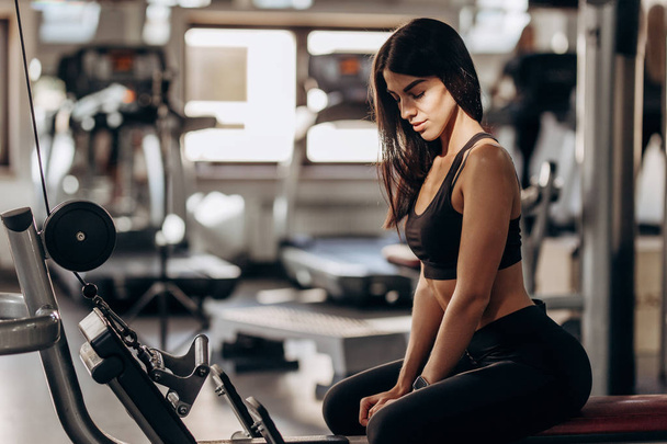 Athletic girl dressed in black sports top and tights is sitting on the bench in the gym - Fotoğraf, Görsel