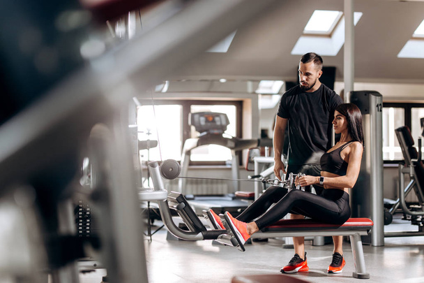Beautiful slim girl dressed in black sport clothes is doing exercises for the abdominals  on a special exercise machine under the supervision of a coach in the gym - Foto, immagini