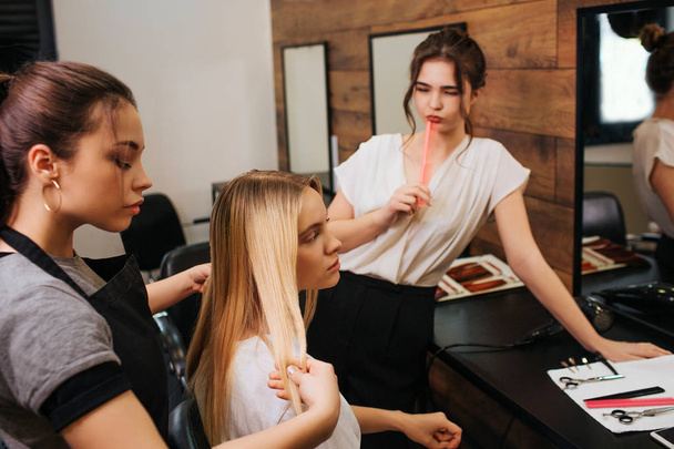 Молода жінка-перукар з чорним фартухом уважно дивиться на нитку світлого волосся, тримаючи її перед професійними процедурами догляду за волоссям в салоні
 - Фото, зображення