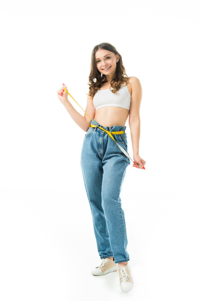 smiling slim girl holding measuring tape on big jeans isolated on white, lose weight concept - Foto, Imagen