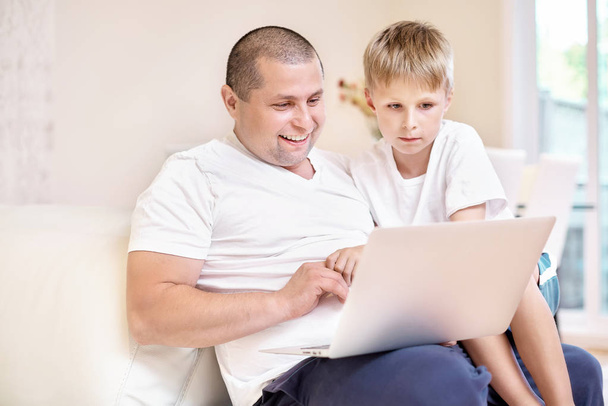 The son and his father are sitting on the couch, looking at a laptop, happy emotions from what he saw, a happy family - Foto, afbeelding