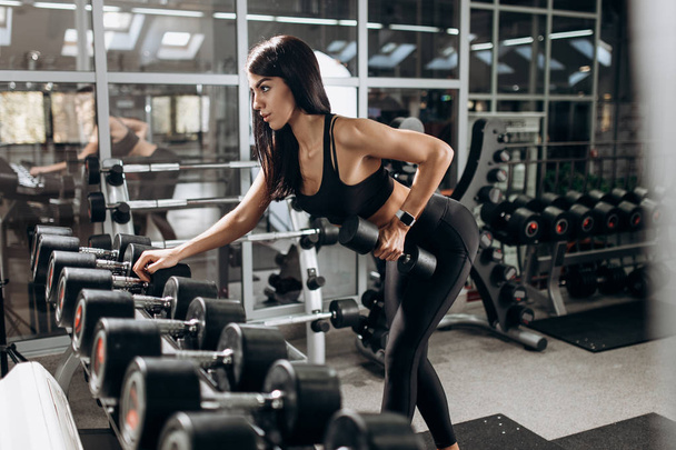 Slim girl dressed in black sport clothes is doing triceps exercise with dumbbell in the gym - Foto, Imagen