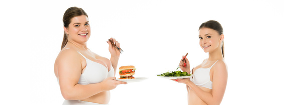 übergewichtige lächelnde Frau in Unterwäsche hält Burger auf Teller, während schlanke, glückliche Frau grüne Spinatblätter isoliert auf weißem Teller isst - Foto, Bild