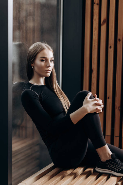 Bella ragazza magra vestita con un abbigliamento sportivo nero è seduta sul davanzale in legno nella moderna palestra
 - Foto, immagini