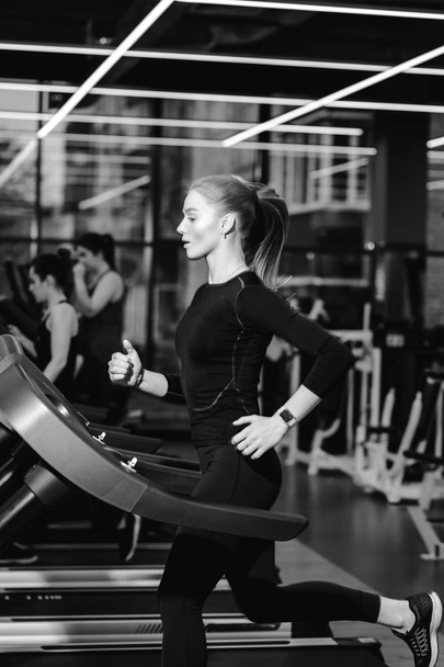 Black and white photo of the athletic girl dressed in a black sportswear running on the treadmill in the modern gym - 写真・画像