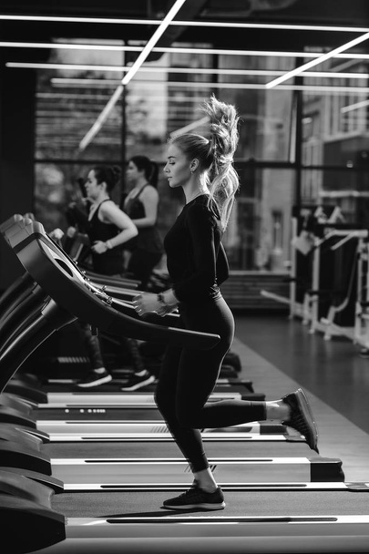 Black and white photo of the athletic girl dressed in a black sportswear running on the treadmill in the modern gym - Valokuva, kuva