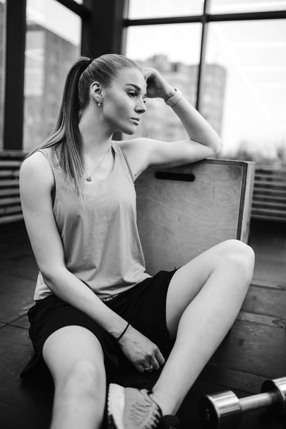 Black and white photo of the athletic girl dressed sport shorts and t-shirt is sitting on the floor next to the box in the modern gym with big windows - Foto, Bild