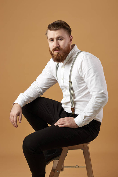 Stylish red-haired man with beard dressed in a white shirt and black trousers with suspender sits on a wooden stool on a beige background - Фото, зображення
