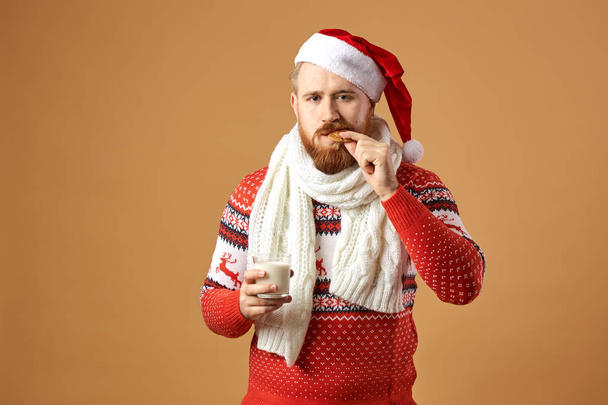 Red-haired man with beard dressed in a red and white sweater with deer, white knitted scarf and a hat of Santa Claus holds a glass of milk and bites a cookie on a beige background - Photo, Image