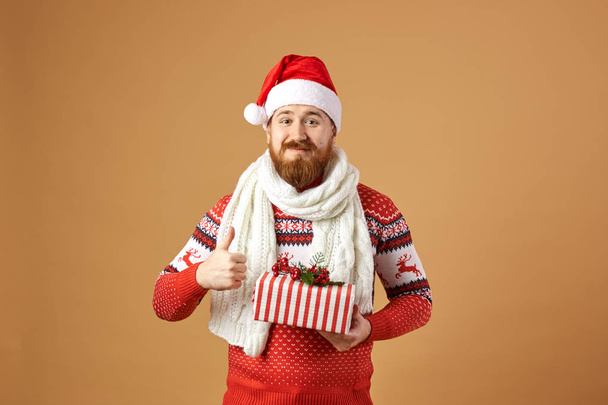 Glad red-haired man with beard dressed in a red and white sweater with deer, white knitted scarf and a hat of Santa Claus holds a Chistmas present in his hands on a beige background - Φωτογραφία, εικόνα