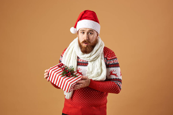 Surprised red-haired man with beard dressed in a red and white sweater with deer, white knitted scarf and a hat of Santa Claus holds a Christmas present in his hands on a beige background - Valokuva, kuva