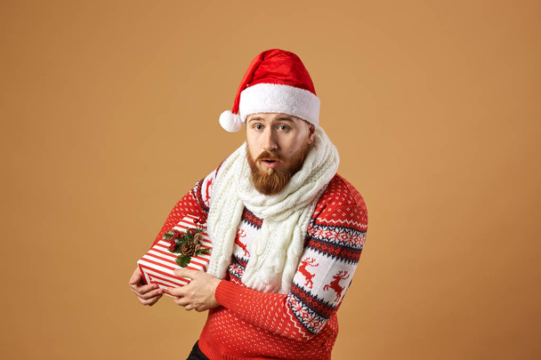 Red-haired man with beard dressed in a red and white sweater with deer, white knitted scarf and a hat of Santa Claus holds a Christmas present in his hands on a beige background - Φωτογραφία, εικόνα