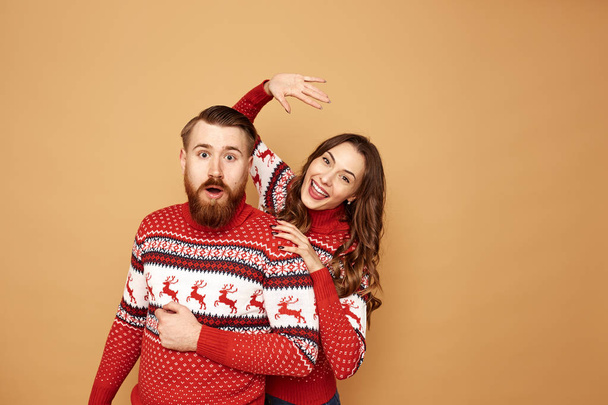 Funny girl and a guy dressed in red and white sweaters with deer stand together on a beige background in the studio - Photo, image