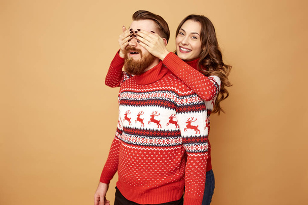 Girl closes the guys eyes with her hands on a beige background in the studio.They both dressed in red and white sweaters with deer. - Photo, Image