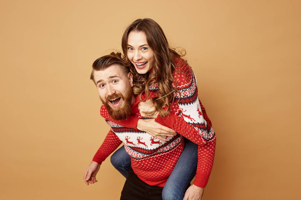 Guy is holding on his back girl and both dressed in red and white sweaters with deer in the studio on a beige background - Foto, afbeelding