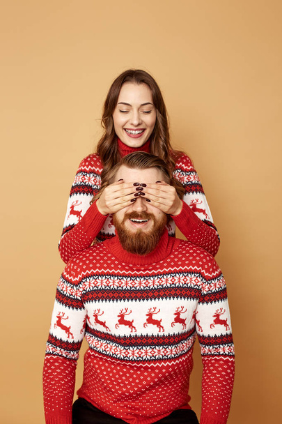 Girl closes the guys eyes with her hands on a beige background in the studio.They both dressed in red and white sweaters with deer. - Foto, Imagen