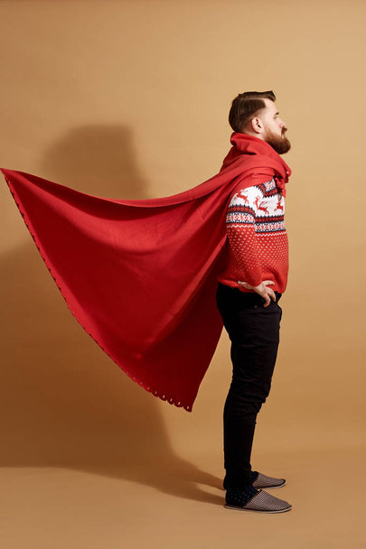 Red-haired man with beard dressed in a red and white sweater with deer and red cape  fluttering in the wind and sleepers stands on a beige background - Fotó, kép