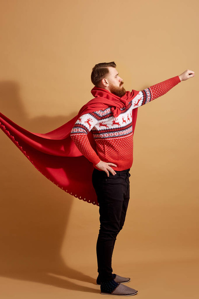 Red-haired man with beard dressed in a red and white sweater with deer and red cape  fluttering in the wind and sleepers stands on a beige background - Photo, image