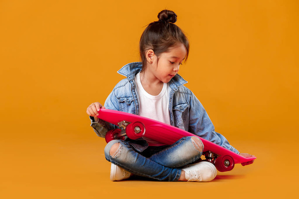 Stylish little child girl with skateboard in denim on orange background - Foto, immagini