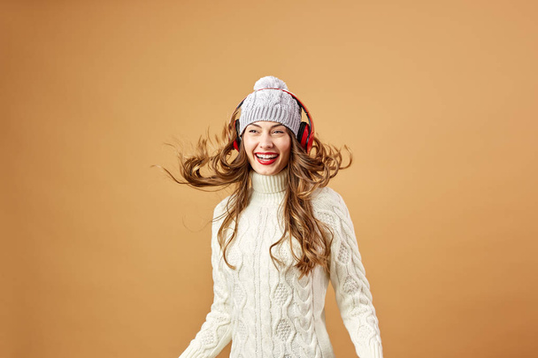 Glad girl in red headphones dressed in white knitted sweater and hat is having fun on a beige background in the studio - Fotoğraf, Görsel