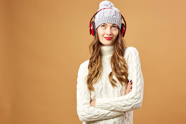 Nice girl in red headphones dressed in white knitted sweater and hat stands on a beige background in the studio - Fotoğraf, Görsel